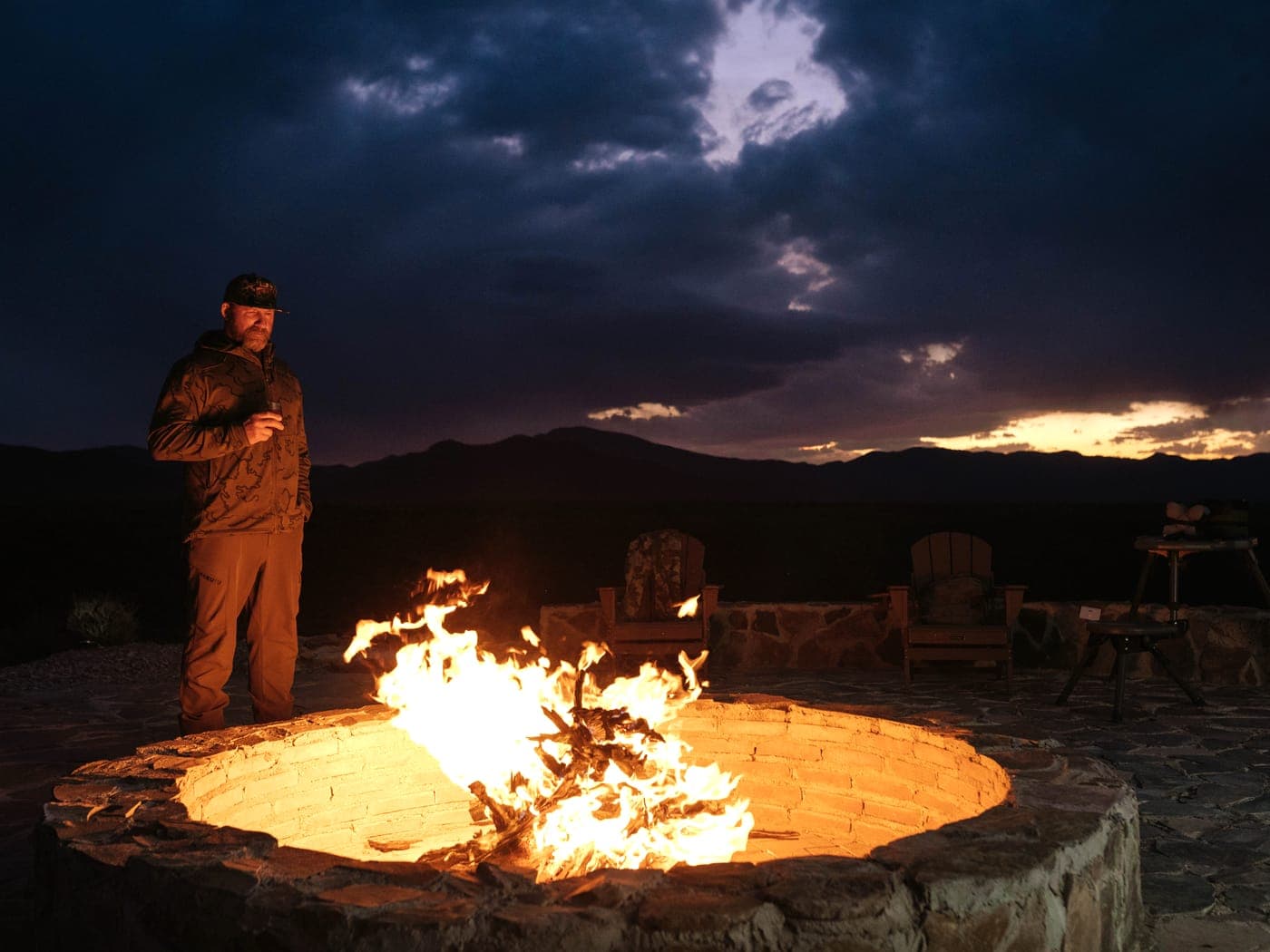 A guest beside a blazing stone fire pit at blue hour, the mountains behind